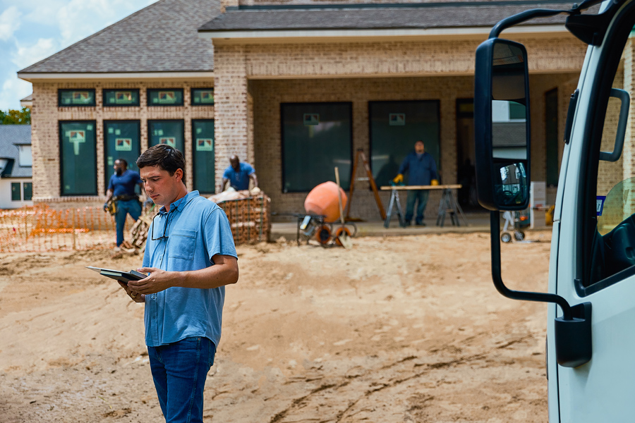 A man with a tablet stands on a construction site where windows are being installed in a new construction home.