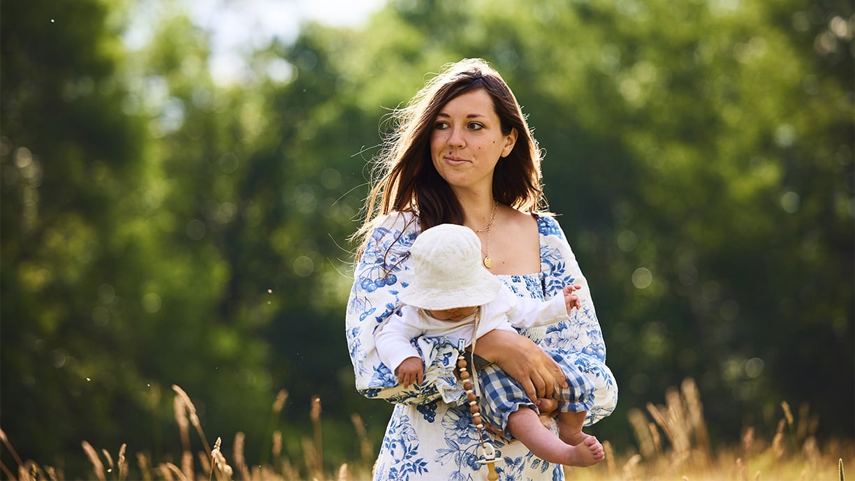 A woman wearing a white dress with blue flowers carries a baby wearing a white bucket hat as she walks across a field of golden grass with a woods behind her.