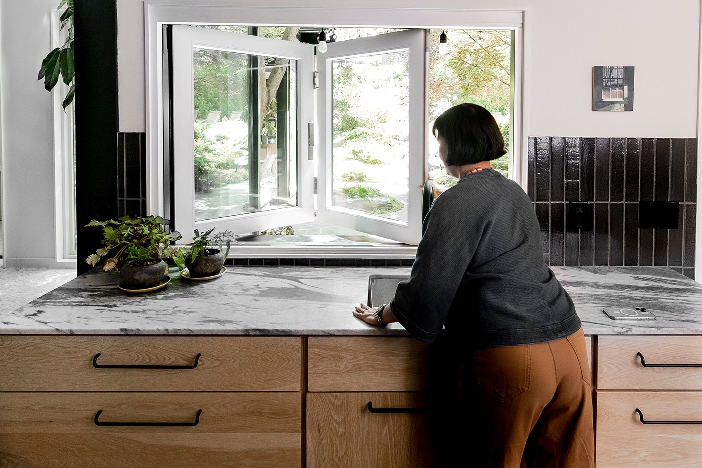 A woman opens a Pass-Through window above a kitchen sink, marble countertops, and potted plants.