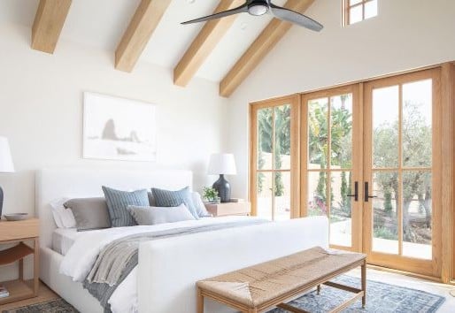 A bedroom with a white bedspread and gray-toned pillows, wooden ceiling beams, and hinged outswing French doors with sidelights and specified equal light grilles.