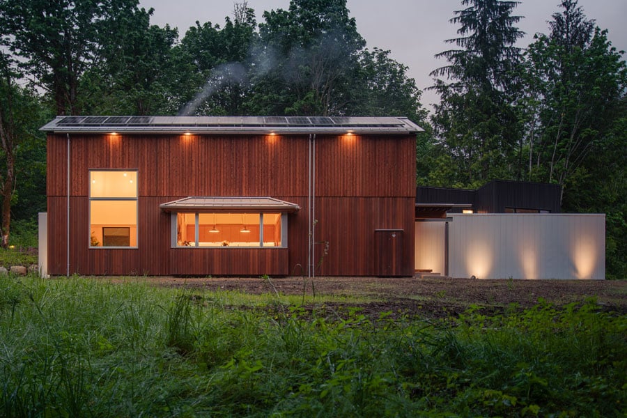 A barn-shaped home clad in vertical cedar with large-scale windows and a solar array on the roof is surrounded by meadow and forest.