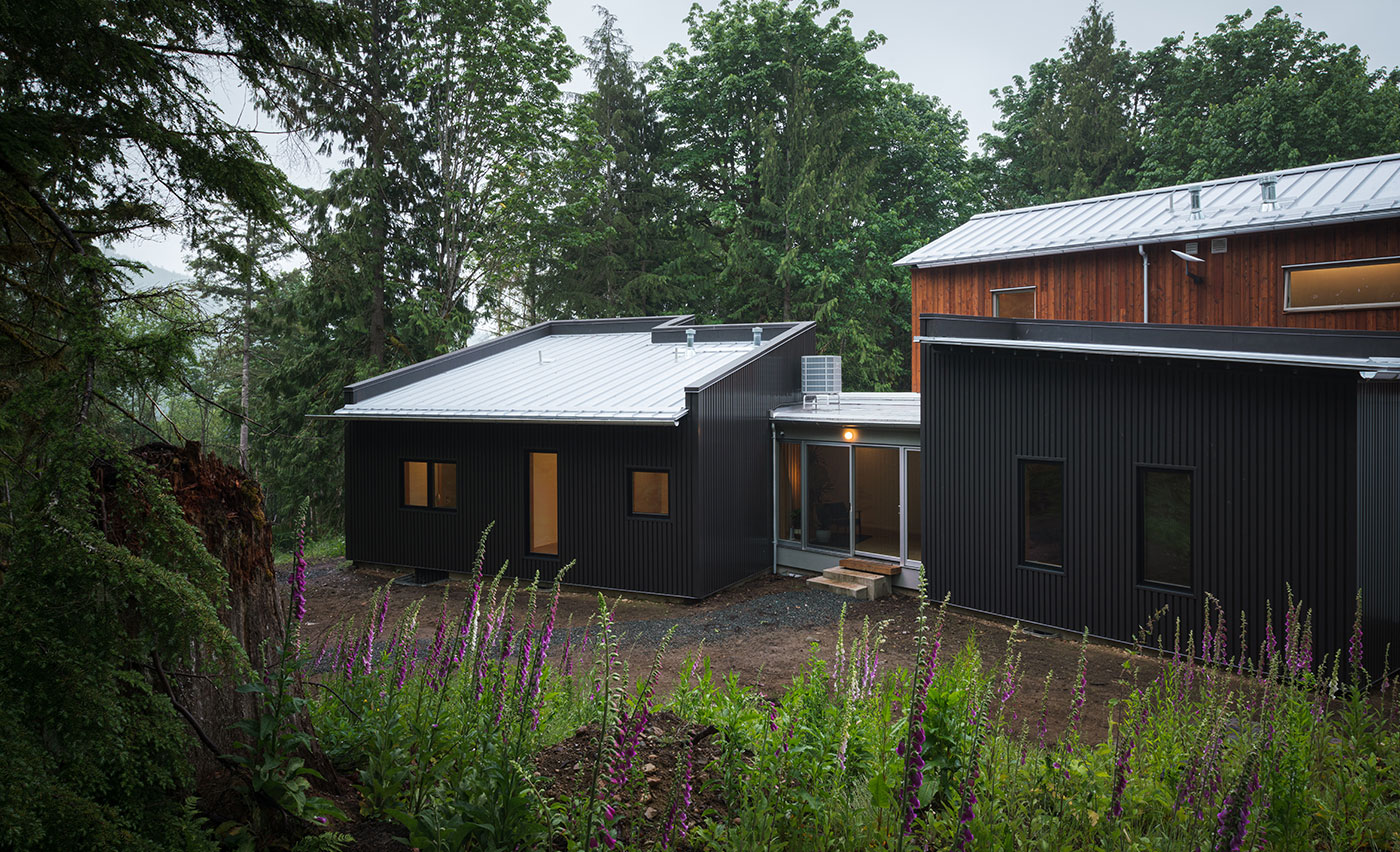 An exterior shot taken from a small hill overlooking a home composed of various volumes, including two clad in vertical black metal that are connected via a glass hallway and another volume that’s clad in cedar behind.