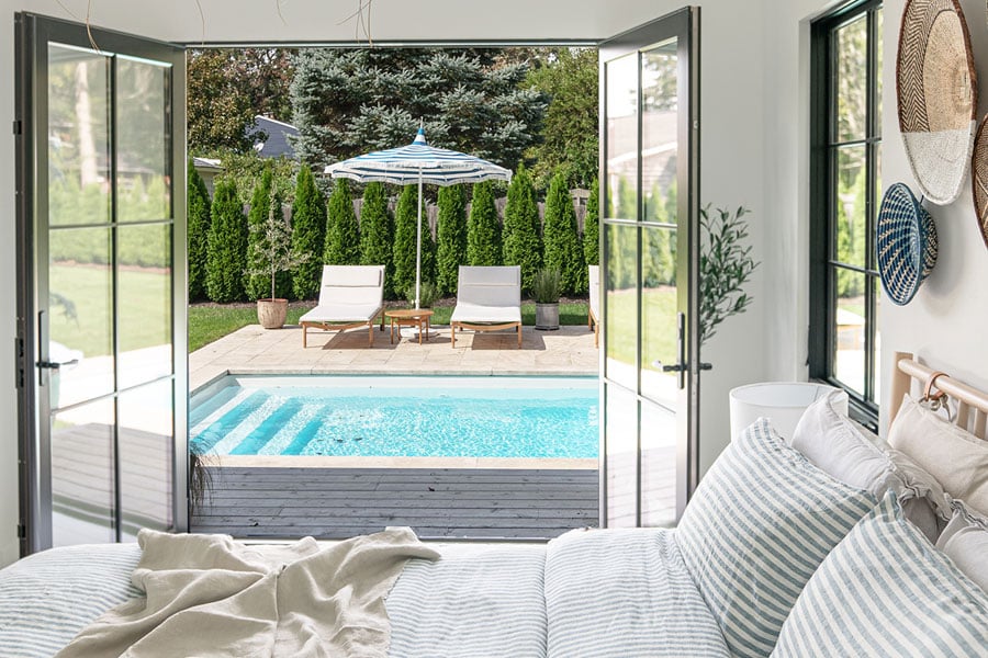 Two interior shots of a bedroom featuring a set of black E-Series Hinged Patio Doors that opens up to a deck and swimming pool outside.