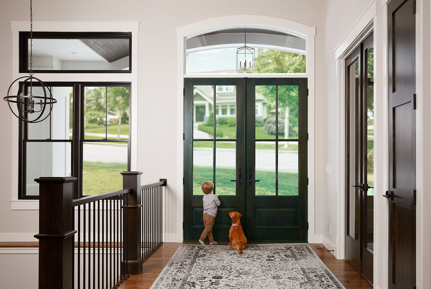 A small child and dog stand in a front hall with wood floors and a rug and look outside through a set of double front doors.