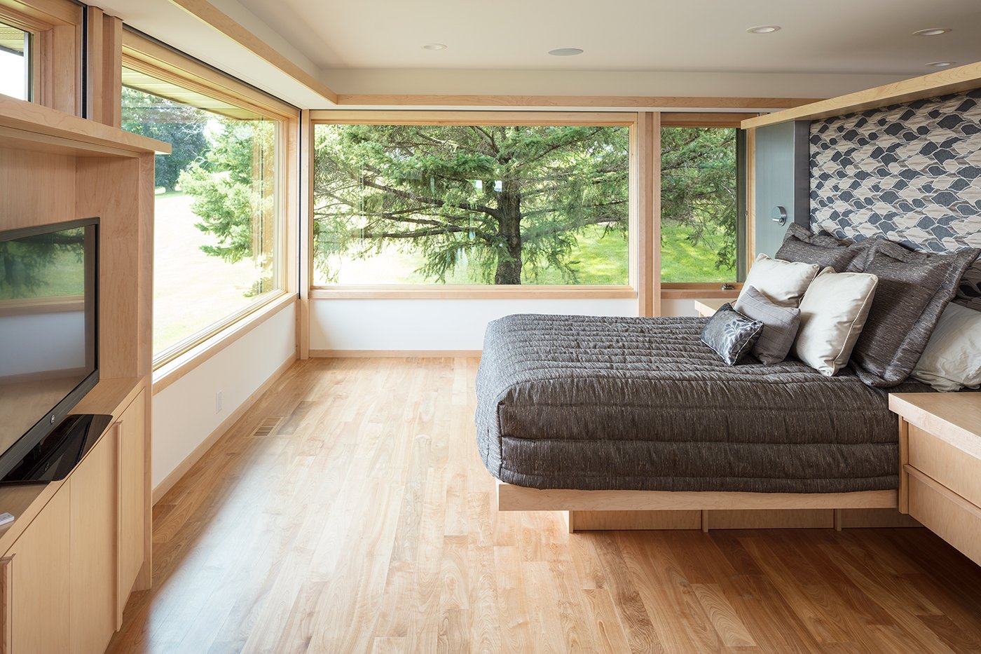 A bedroom with a platform bed surrounded by two walls of Andersen® A-Series Picture and Casement Windows with maple interiors.