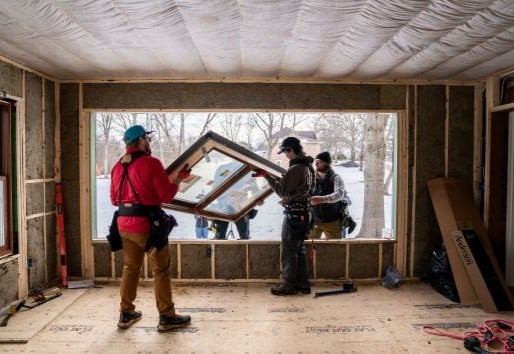 Four men install a window in a large opening of a home with snow outside.