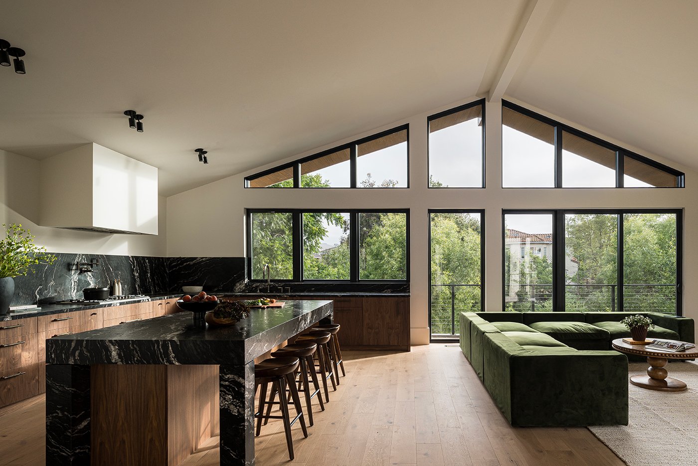 An open living room and kitchen layout with hardwood floors, black marble countertops, and a plush green couch with a wall of 100 Series Windows and Gliding Patio Doors, and transom windows with black frames. 