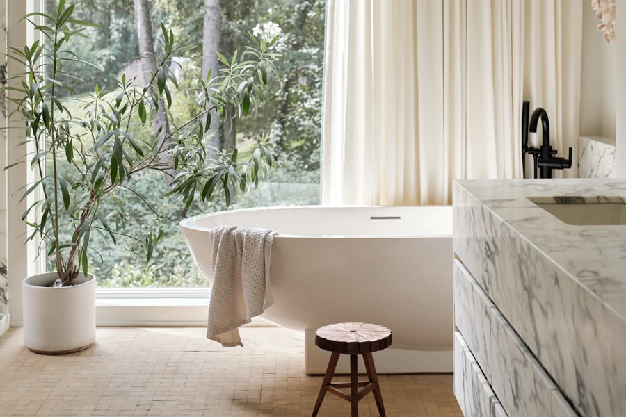 A bathroom with a large soaking tub in front of a floor-to-ceiling window that’s half covered by a white curtain and a floating vanity made of blue-veined marble.