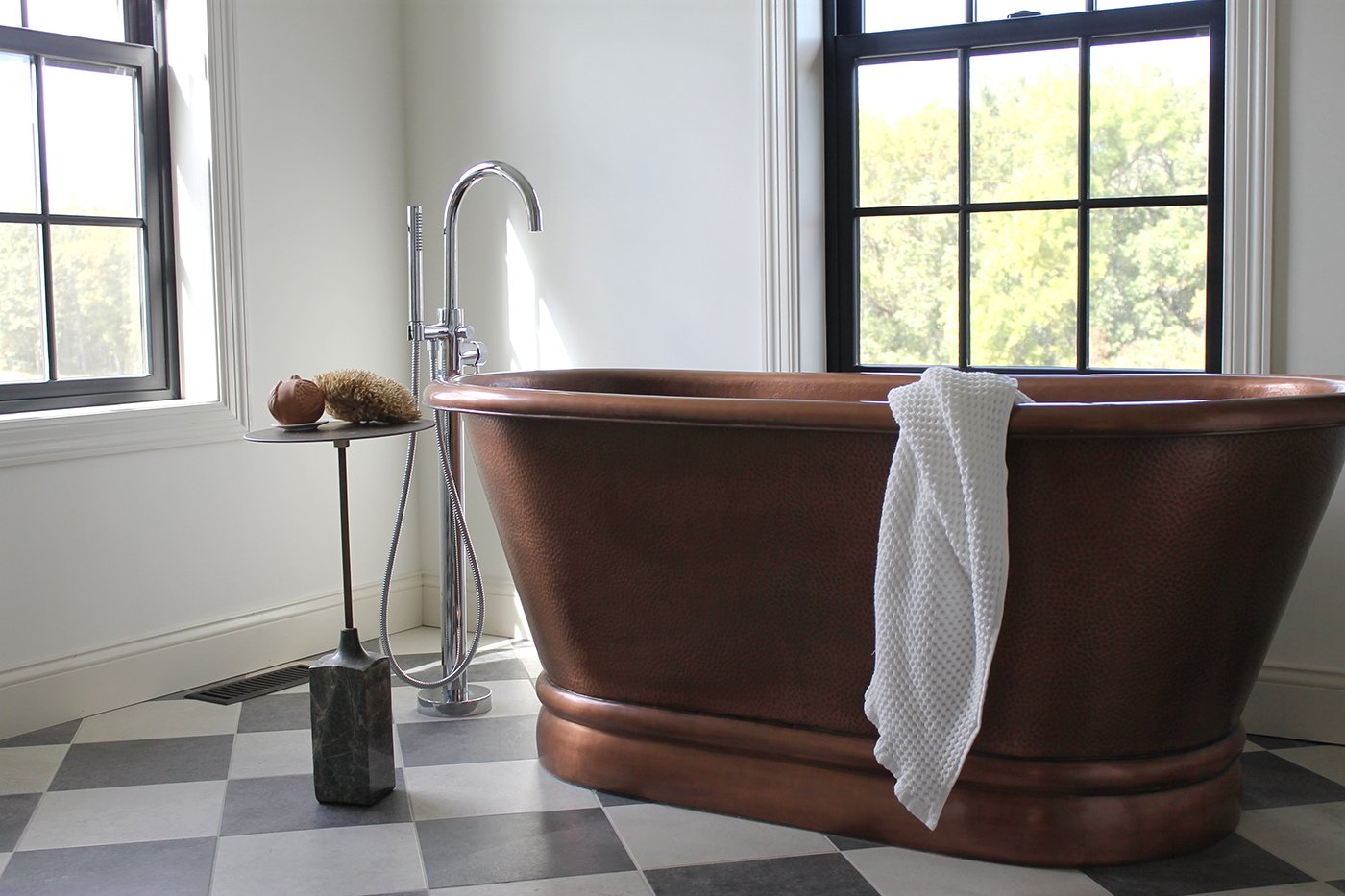Two double-hung windows bring natural light into a bathroom with a copper tub and black-and-white checkerboard floor.