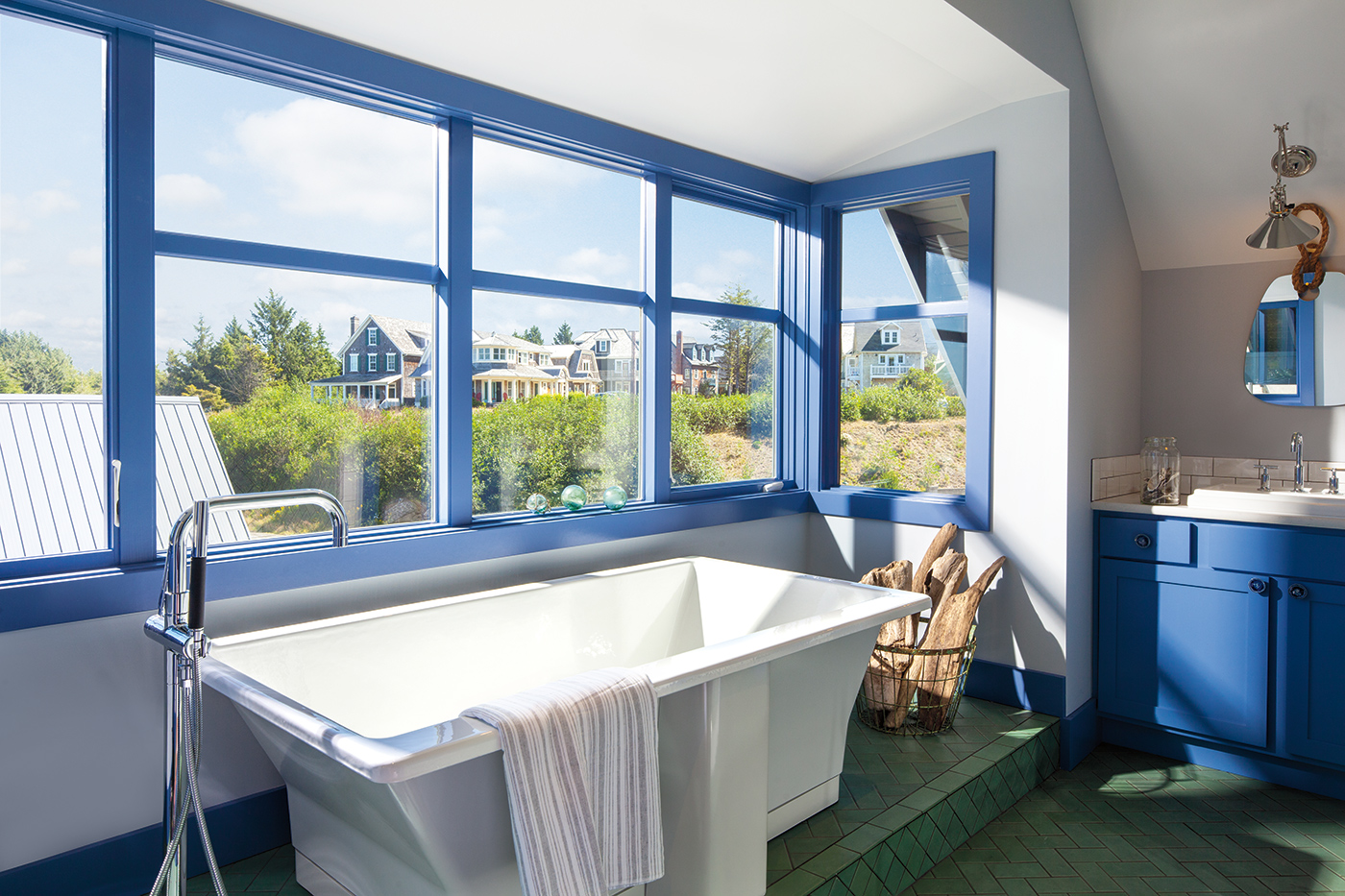 A bathtub surrounded by periwinkle blue windows and window trim with views of a hill and shingle-sided homes.