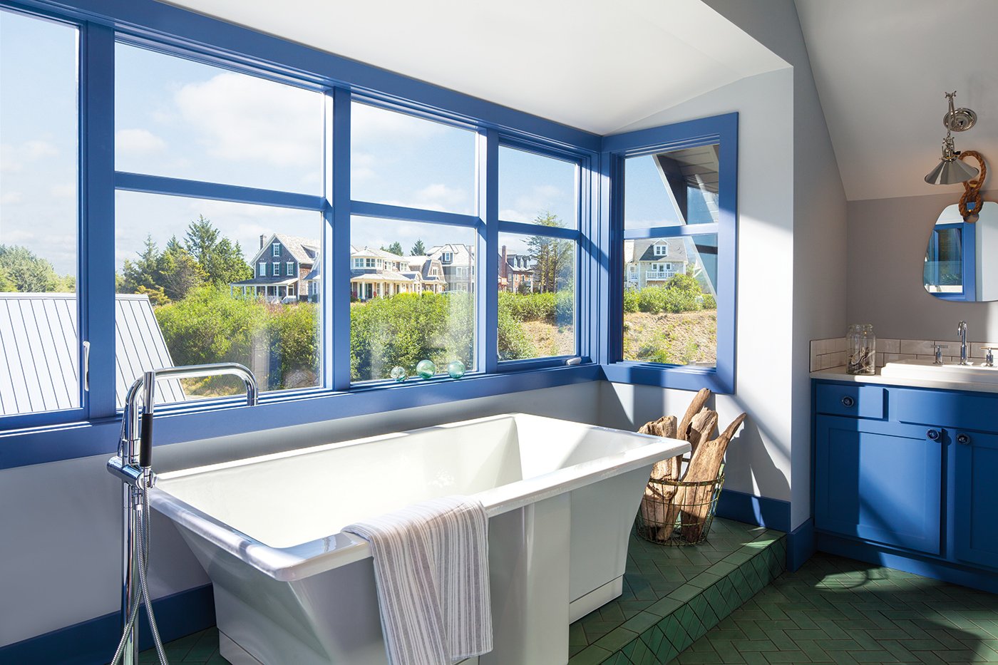A bathtub surrounded by periwinkle blue windows and window trim with views of a hill and shingle-sided homes.