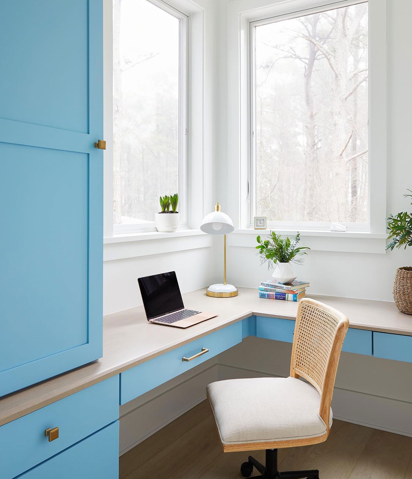 A home office with blue painted cabinetry, a white oak work surface, and casement corner windows that make the space airy and bright.