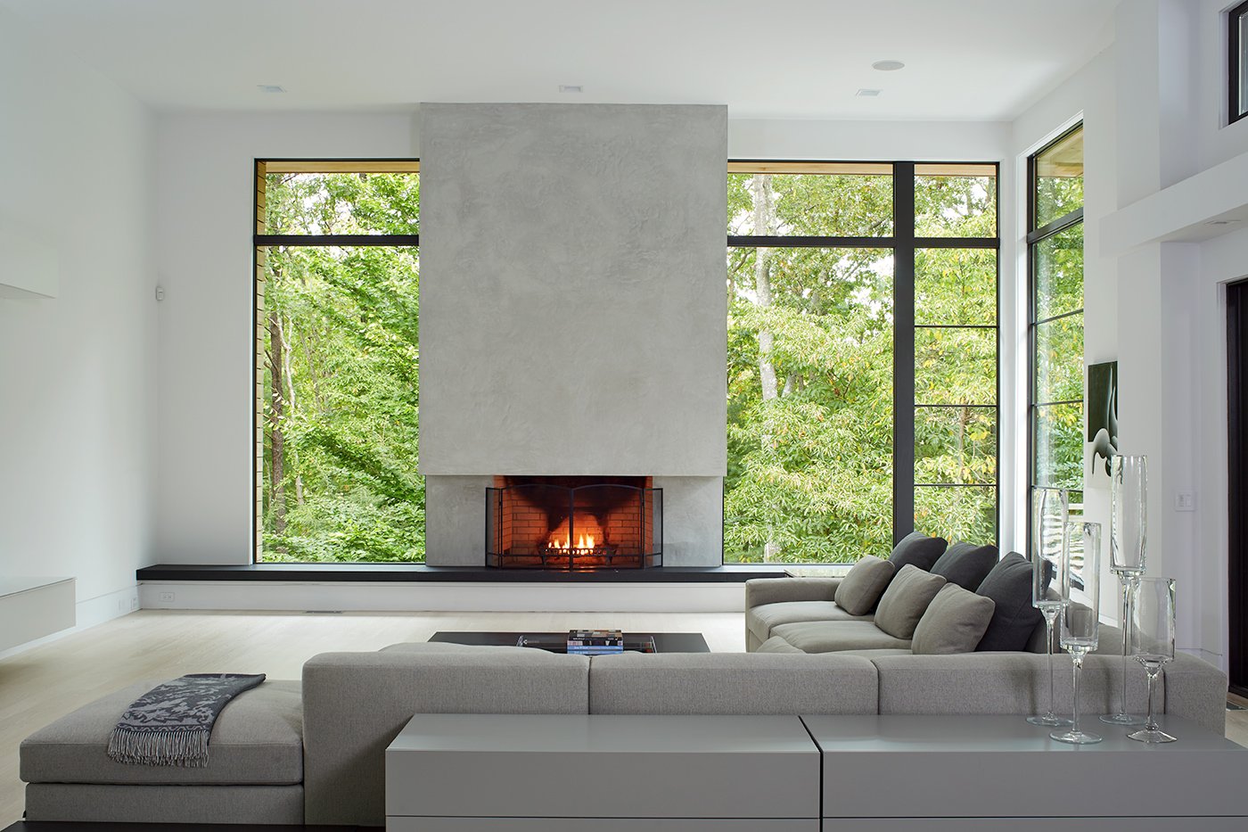 A living room in a modern home that features a concrete fireplace surrounded by floor-to-ceiling windows with black frames.
