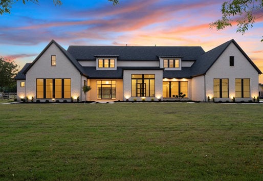 large, ranch style modern farmhouse nestled on a large, well-manicured green lawn. The exterior is a warm white brick color with large black windows with straight line grilles and a black roof that houses three large gables