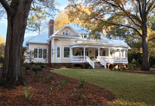 A yellow Victorian home sitting on a large lawn with a traditional, southern front porch. The home has white trim around the porch and all white windows. The windows feature diamond grille patterns.