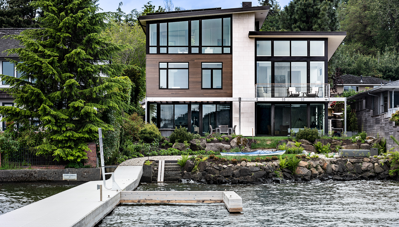 A home next to the water with a white dock, with flat roofs and large windows and patio doors with black frames.