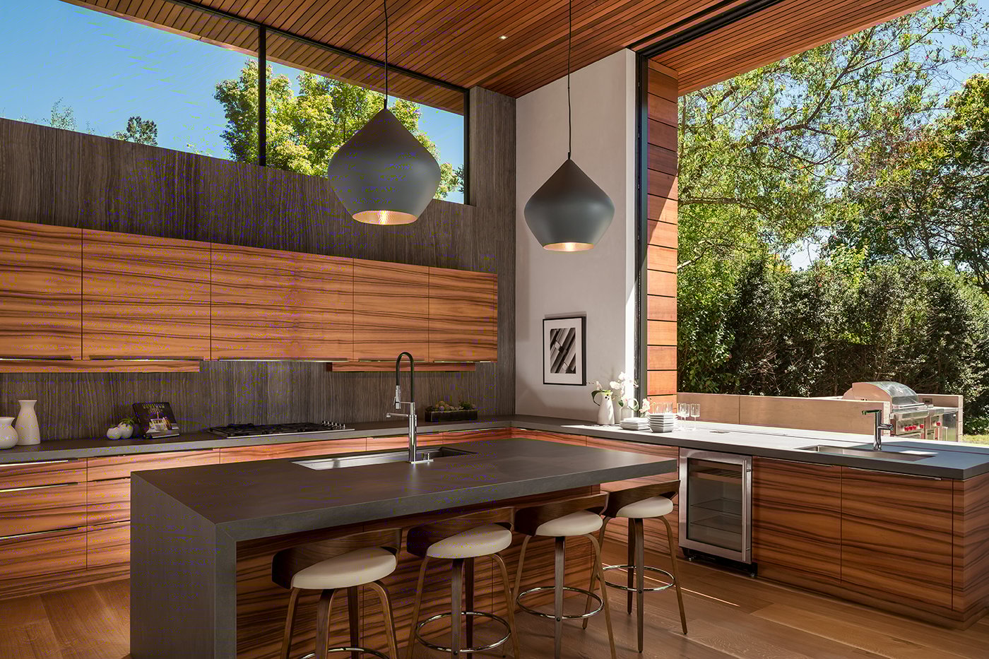 A kitchen with a modern design featuring wood cabinetry, black clerestory windows, and a monumental pass-through window that’s installed flush with the countertop.