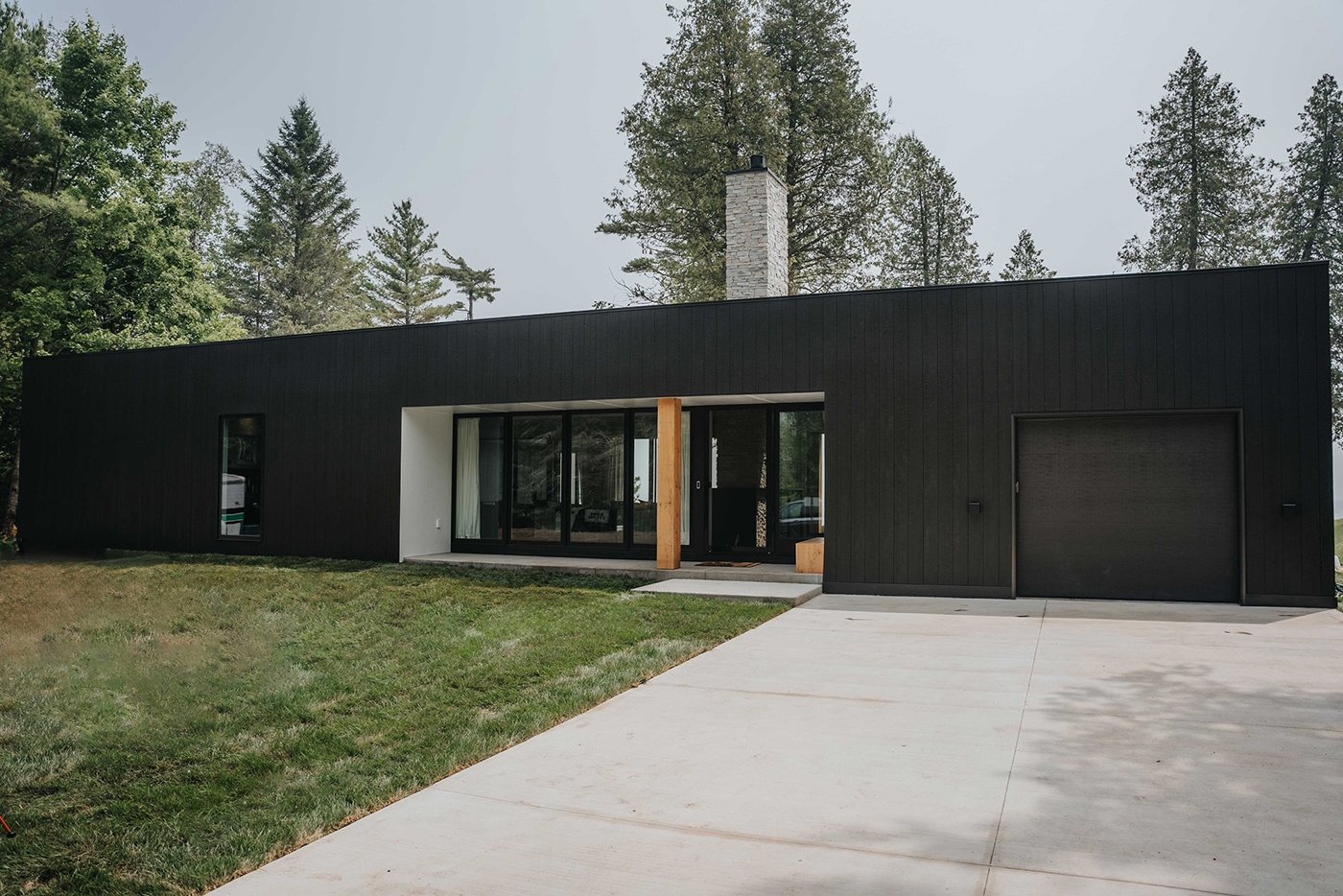 A modern black home with a flat roof, grey brick chimney and wooden pillar in front of a sliding glass door and tall windows set back in an entry porch. 