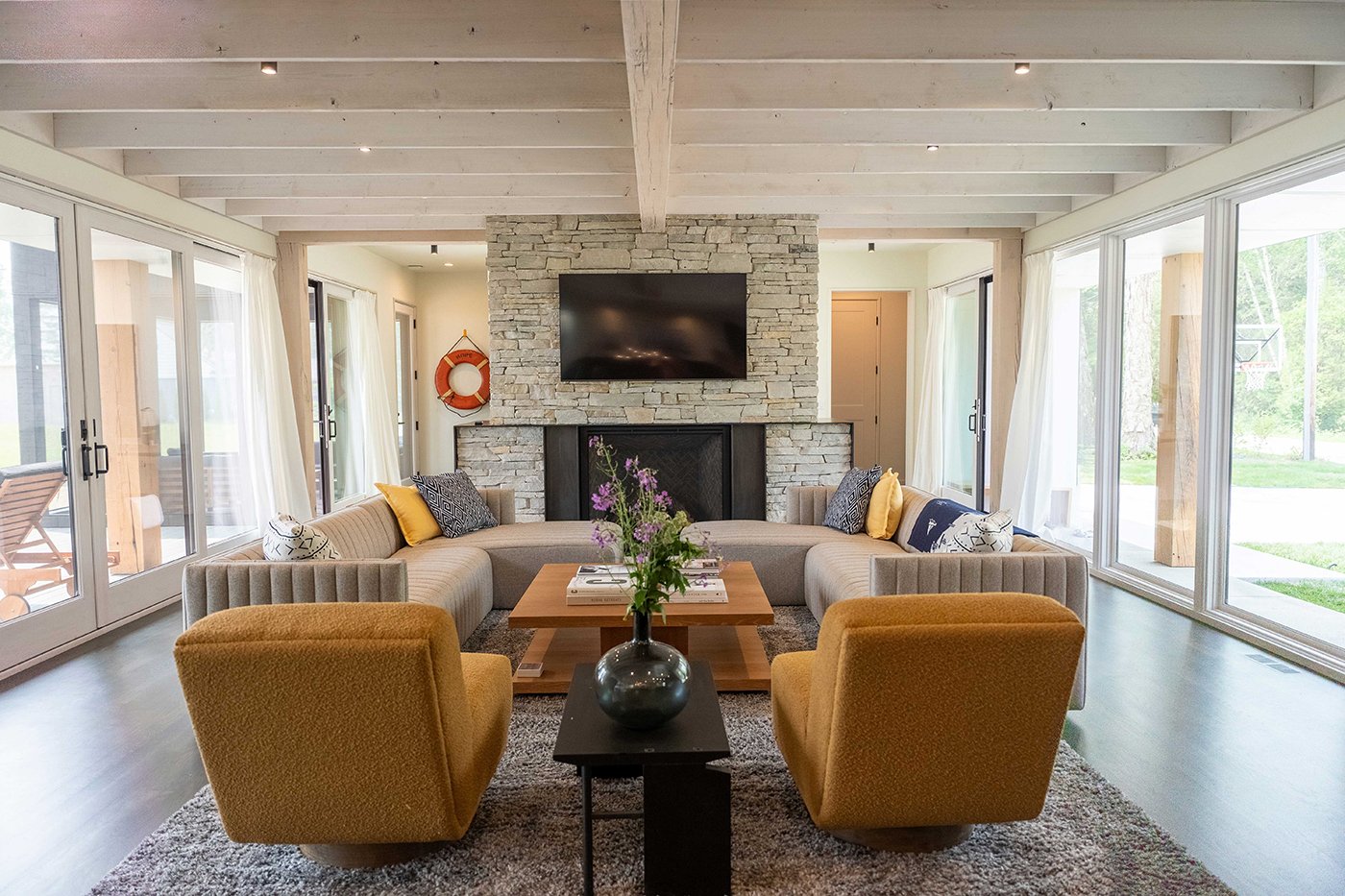 A living room with a stone fireplace, couches and chairs, and entire walls of windows and patio doors opposite each other. 