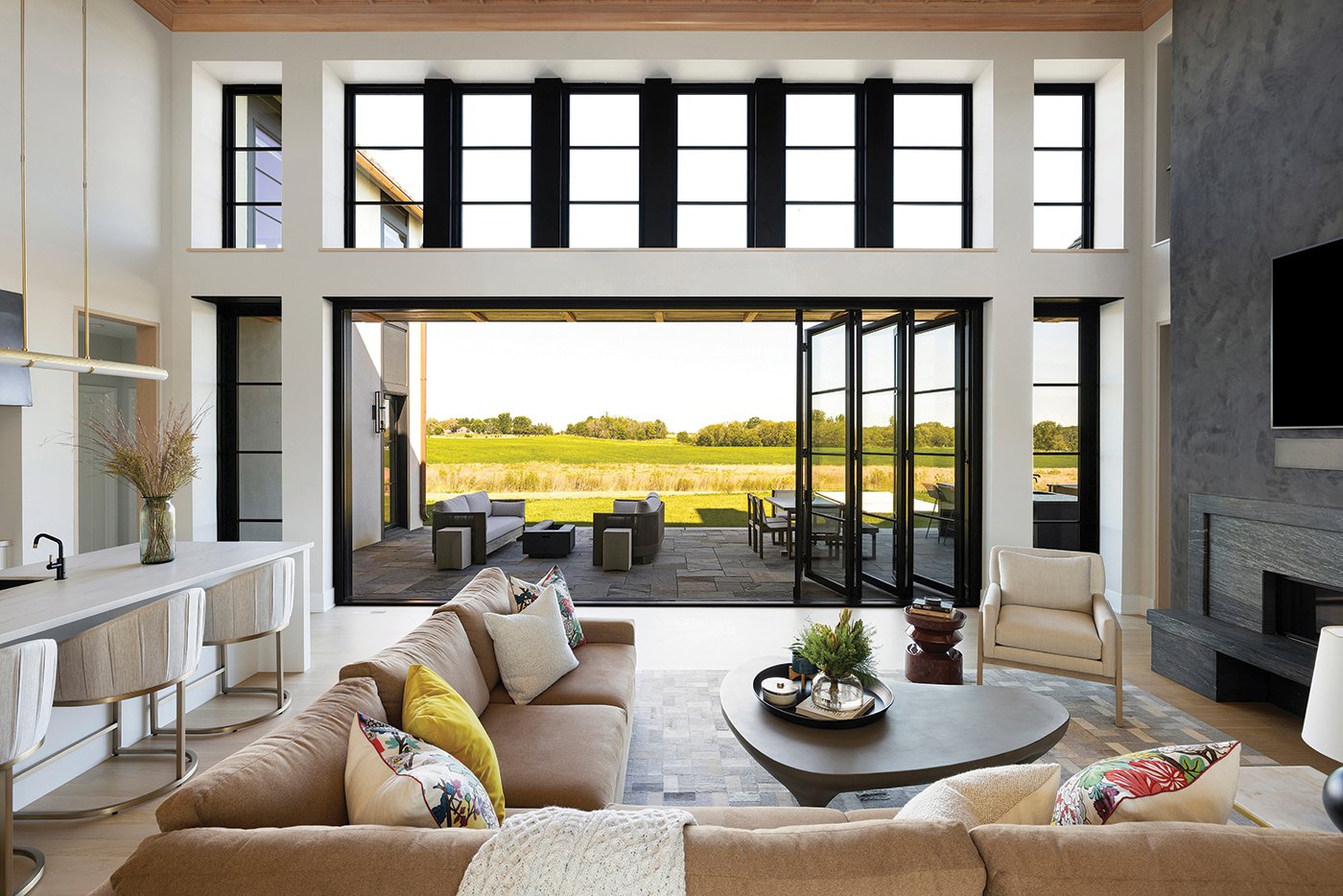 A modern Tudor home with tall ceilings and an expansive, black Andersen Folding Outswing Door folding open to reveal an outdoor lounge and dining area outside with an expansive prairie view. 