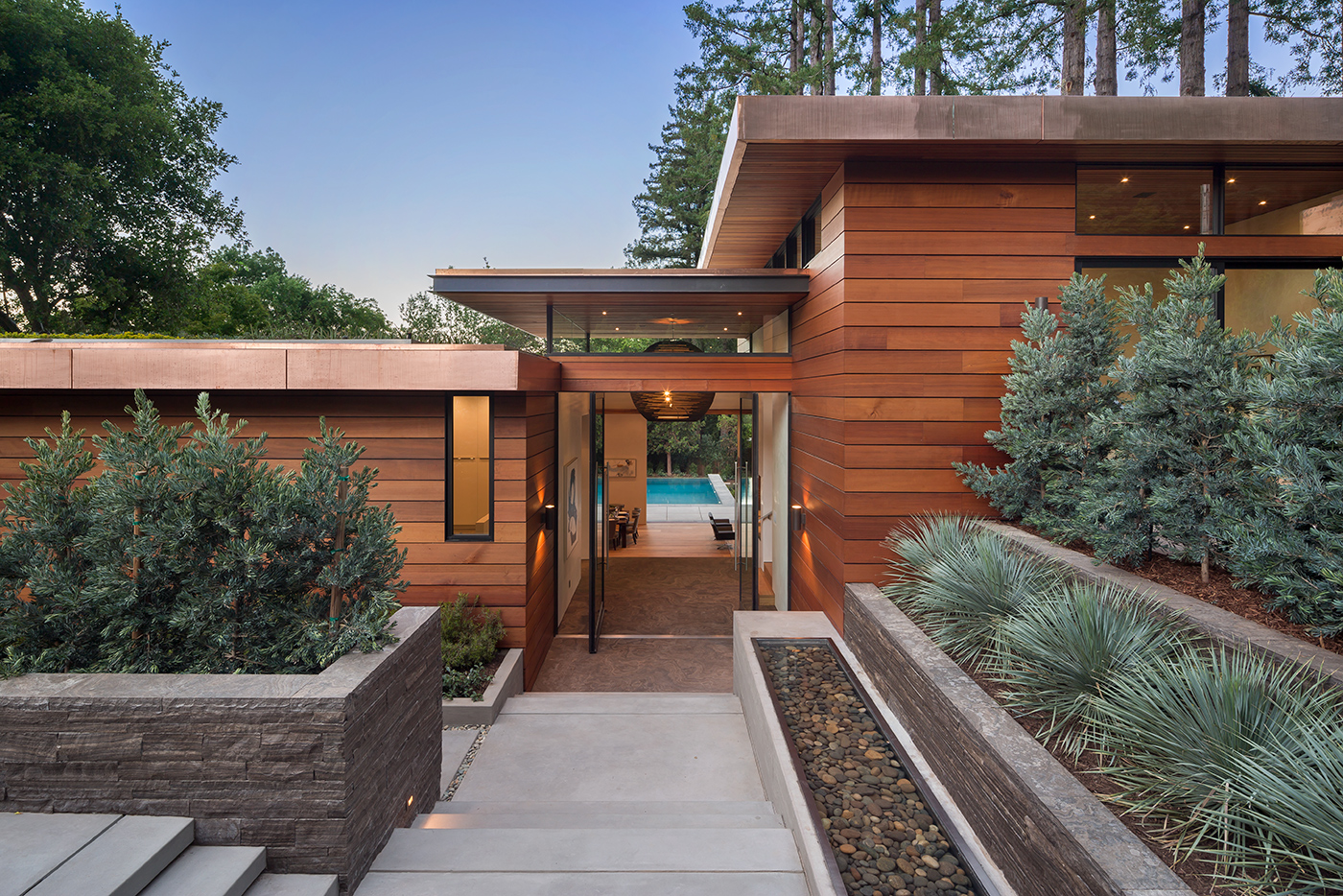 The exterior of a mahogany-colored California home with a double Andersen Pivot Door in the center allowing for a view all the way through the home to the pool and Redwoods beyond. 