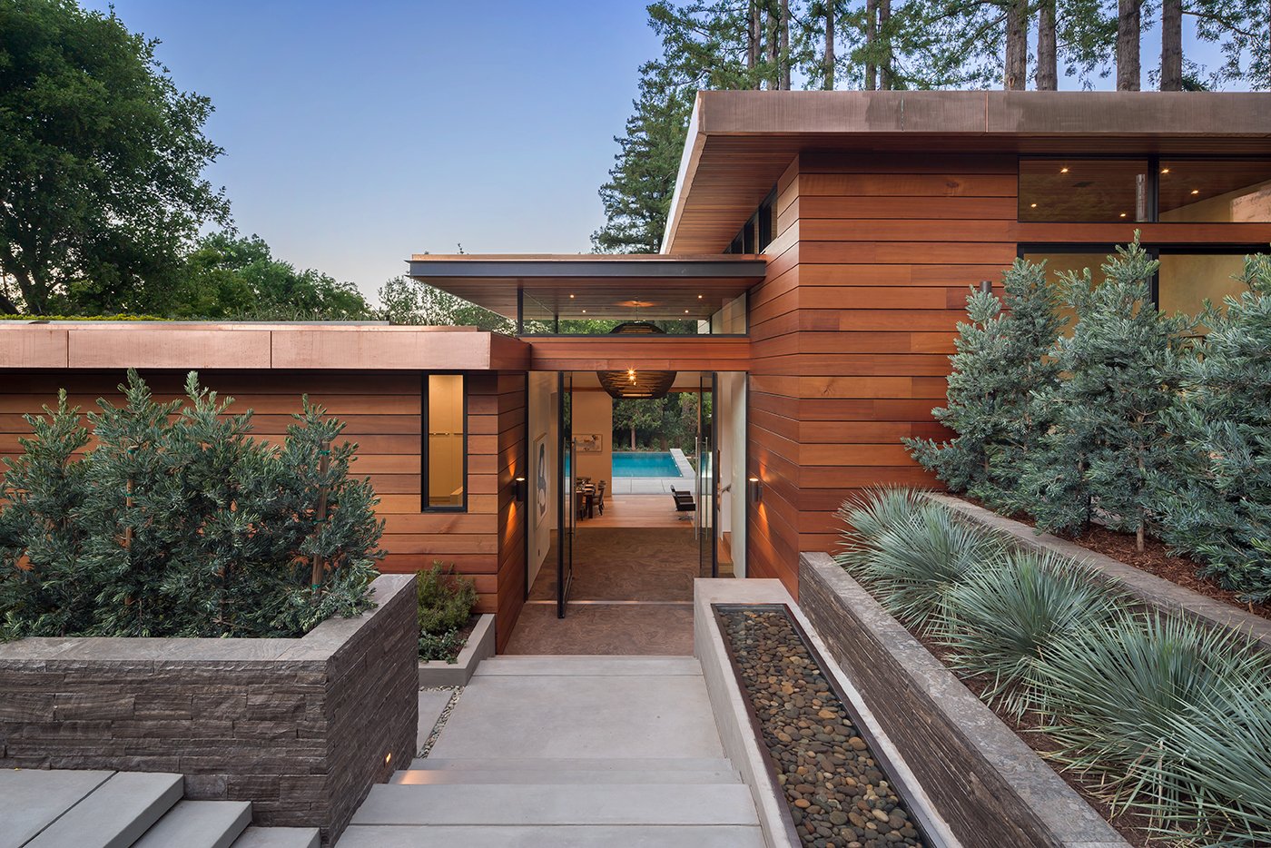 The exterior of a mahogany-colored California home with a double Andersen Pivot Door in the center allowing for a view all the way through the home to the pool and Redwoods beyond.