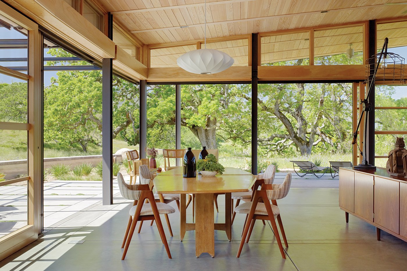 A modern, natural open-air dining room with corner and two side walls open to the sunny treescape and free-flowing fresh air by way of an Andersen Liftslide Door that meets at the corner with an interior wood finish. 