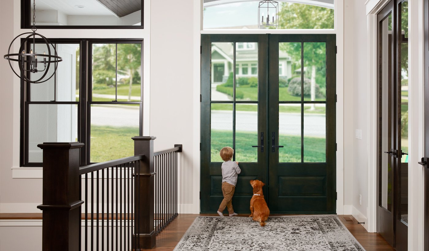 Interior view of a child and dog peering out of the large glass panes of a black double front door next to black windows, white walls, and transom window. 