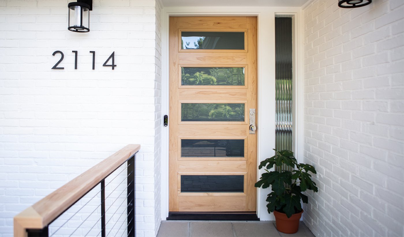 A modern front door with a light-colored wood exterior and horizontal glass panels popping against the white brick walls of a Mid-Century Modern home.