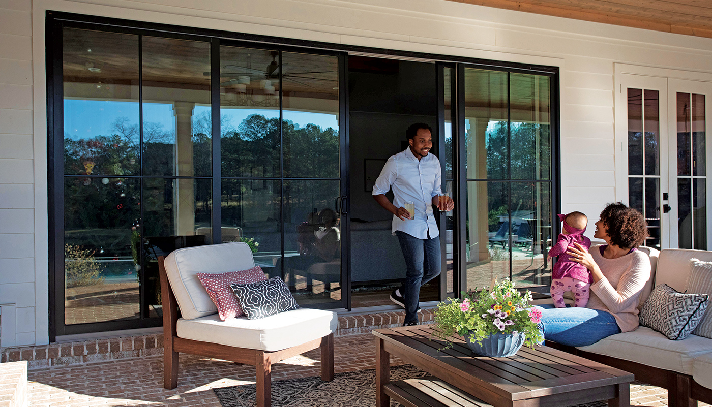 A man walks out an Andersen 400 Series Gliding Patio Door to a patio where a woman and baby are seated on a couch.