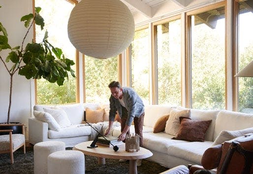 A man arranges books on a coffee table next to a white couch in a living room that has floor-to-ceiling picture windows with white oak frames.