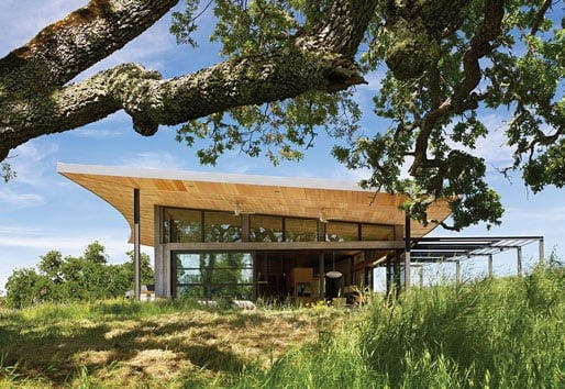 An exterior view of a modern ranch that showcases its generous roof overhang and sunshade system along with the surrounding landscape of rolling hills and mature oak trees.