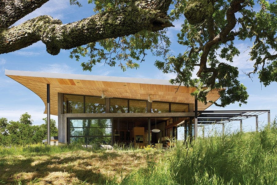 An exterior view of a modern ranch that showcases its generous roof overhang and sunshade system along with the surrounding landscape of rolling hills and mature oak trees.
