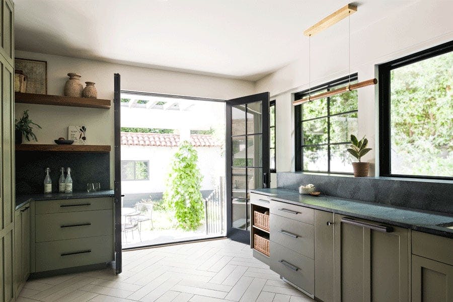 A kitchen with French doors that open to the backyard and windows above the kitchen cabinetry offering more views of the greenery outside.